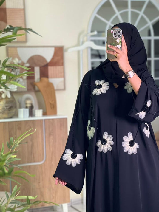 Model wearing a black abaya with white floral patterns, taking a mirror selfie.